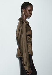 Brown satin blouse with a structured collar, gathered waist, long sleeves, and buttoned cuffs, paired with black pants. Model facing side.