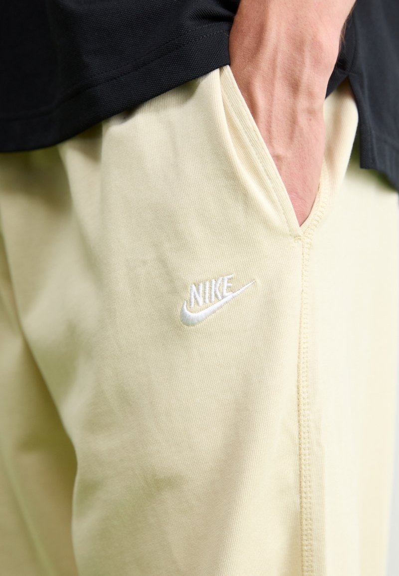 Jasnożółte spodnie Nike z teksturowanej tkaniny. Posiadają białe haftowane logo na kieszeni, widoczne szwy oraz luźny krój.