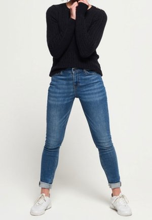 Person står med benene spredt, iført opsmøgede blå jeans, hvide sneakers og en sort langærmet sweater, hænder nær ansigtet.