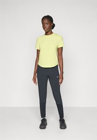 Helles gelbes, kurzärmliges Sportshirt mit Rundhalsausschnitt, kombiniert mit schwarzen, schmal zulaufenden Sporthosen. Beide Teile sind mit Nike-Logos versehen.