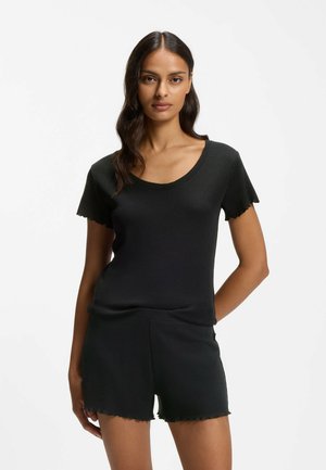 Femme aux longs cheveux ondulés portant un haut noir à manches courtes et à côtes, avec un short noir assorti, debout devant un fond blanc uni.