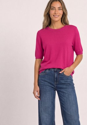 Femme aux cheveux châtain clair portant un haut rose vif à manches courtes et un jean bleu taille haute, souriante avec une main dans la poche.