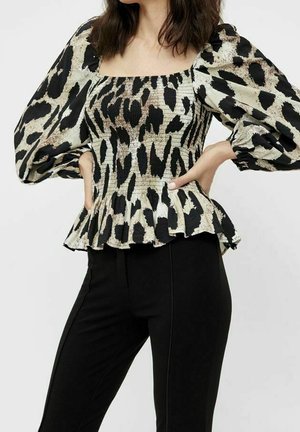Frau trägt eine Bluse mit Leopardenmuster und Puffärmeln, einem gerafften Oberteil und Schößchen, kombiniert mit eng anliegenden schwarzen Hosen. Hände in der Hüfte.