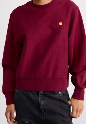 Osoba ubrana w bordową bluzę Carhartt z długimi rękawami i czarne dżinsy, ręce swobodnie wzdłuż ciała.