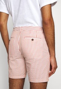 Homme portant un short rayé verticalement blanc et rose et une chemise blanche à manches courtes, vu de dos de la taille jusqu’au milieu de la cuisse.