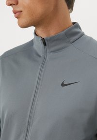Chaqueta gris con cremallera hecha de tejido texturizado, con un sutil logo negro de Nike en el lado izquierdo del pecho.