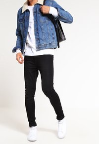 Farkkutakki, jossa on kermainen sherpa-kaulus, valkoinen collegepaita, mustat tiukat farkut ja valkoiset lenkkarit. Asusteessa on rento tyyli.