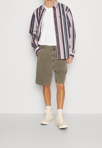 Man in een witte T-shirt, open gestreepte overhemd, olijfgroene shorts, witte sokken en crème hoge sneakers, staand met één hand in zijn zak.