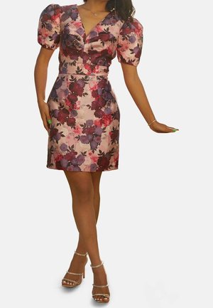 Kobieta w krótkiej kwiecistej sukience z bufkami na rękawach, dekoltem w serek i szpilkach na paskach ozdobionych perłami, pozująca z jedną wyciągniętą ręką.