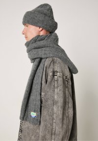 Grå uldebrætte og tørklæde, begge med en blød tekstur. Tørklædet svøber sig om halsen og komplementerer en denimjakke med mærkedetaljer.