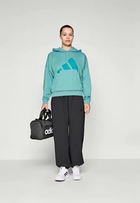 Tirkiz dukserica s kapuljačom i prednjim džepom, s plavim naglaskom, uparen s labavim crnim hlačama i bijelim tenisicama, drži crnu Adidas torbu.