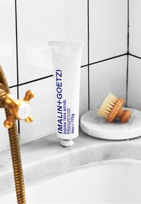 Weißer Tube des Malin+Goetz Jojoba-Gesichtspeelings mit blauer Schrift, auf einer Marmoroberfläche neben einer Holzbürste platziert, vor einem gefliesten Hintergrund.