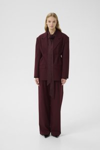 Burgundy tailored blazer with shoulder pads and a flower brooch, paired with wide-leg trousers in matching fabric, featuring a smooth texture.
