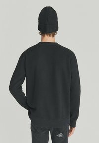Felpa nera con maniche lunghe e colletto a costine, indossata con un berretto lavorato a maglia nero coordinato, vista da dietro.