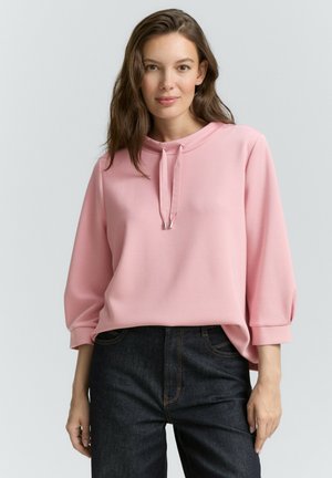 Helles rosa Sweatshirt mit lockerer Passform, ausgestattet mit einem Kordelzugkragen, Dreiviertelärmeln und weichem Material. Kombiniert mit dunklem Denim.