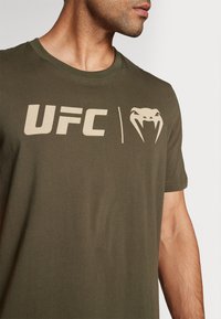 Oliven grøn bomulds t-shirt med et stort beige "UFC" logo og et slangemotiv på venstre side. Korte ærmer og rund halsudskæring.