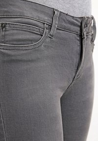 Detailní záběr na šedé džíny, který ukazuje přední kapsu, poutka na pásek a pas na osobě, která má na sobě šedý top.