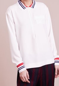 Polo shirt bianco a maniche lunghe con colletto a zip, caratterizzato da accenti a righe blu navy e rosse sul colletto e sui polsini. Tessuto liscio con una vestibilità comoda.