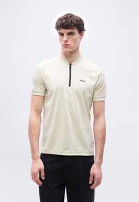 Νεαρός άνδρας φορώντας ένα κρεμ κοντομάνικο πουκάμισο με φερμουάρ και το λογότυπο "BOSS" στο αριστερό στήθος, και μαύρο παντελόνι, στέκεται μπροστά σε απλό λευκό φόντο.