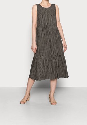 Vrouw draagt een mouwloze zwarte midi-jurk met wit geometrisch patroon en beige sandalen met blokhak, staande op een grijze vloer.