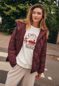 Veste matelassée bordeaux avec capuche portée sur un sweat-shirt blanc orné d'un dessin animé. Un pantalon côtelé beige clair complète la tenue.