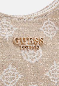 Tissu texturé beige avec des motifs blancs, comportant un logo doré "GUESS EST 1981". Le design inclut des accents délicats et des détails de couture.