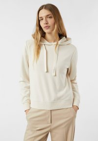 Kremowa bluza z kapturem wykonana z lekkiego materiału, z regulowanym kapturem na sznurek, ściągaczami na rękawach oraz luźnym krojem, zestawiona z beżowymi spodniami.