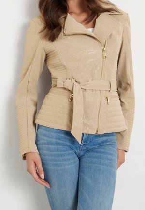 Kvinde iført en beige jakke med bælte, lynlåslommer, asymmetrisk frontlynlås og quiltede detaljer, kombineret med blå jeans.