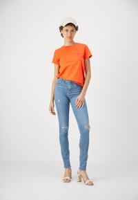 Jasnopomarańczowa bawełniana koszulka z okrągłym dekoltem i krótkim rękawem, zestawiona z jasnoniebieskimi skinny jeansami z dziurami na kolanach i wysokimi obcasami.