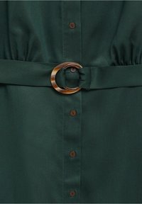 Robe en tissu vert foncé avec des boutons marron et une ceinture assortie dotée d'une boucle ovale en écaille de tortue à la taille.