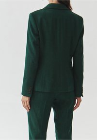 Giacca da completo sartoriale verde scuro con una texture liscia. Dettagli notevoli includono una rever a incavo e polsini con tre bottoni, indossata sopra pantaloni abbinati.
