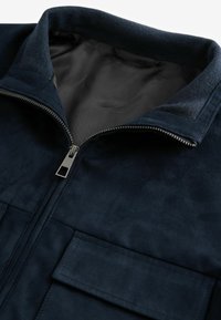 Chaqueta de ante azul marino con cuello alto, cremallera frontal y dos bolsillos en el pecho. Presenta una textura suave y un forro interior en contraste.