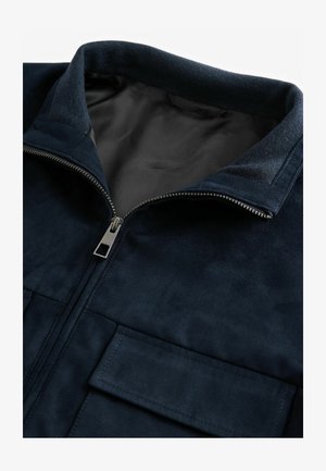 Marineblauwe suède jas met een hoge kraag, een rits aan de voorkant en twee borstzakken. Kenmerkt zich door een soepele textuur en een contrasterende voering.