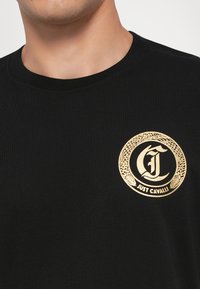 Svart T-skjorte med ribbet halslinning har et sirkulært gull-logo med intrikate detaljer og teksten "JUST CAVALLI" i sentrum.