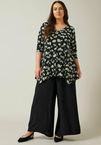 Schwarze Blumentunika mit weißen, grünen und lila Blumen, mit V-Ausschnitt und Dreiviertelärmeln, kombiniert mit weit geschnittenen schwarzen Hosen.