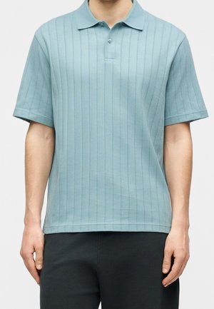 Homme portant un polo bleu clair à manches courtes avec des rayures verticales et un pantalon noir, debout devant un fond uni.
