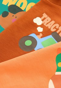 Tre t-shirt di cotone in arancione, marrone e teal con grafiche giocose: un cane, un trattore con fumo e un orso con accenti colorati.