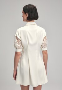 Crème-kleurige jurk met korte pofmouwen van doorschijnende stof en kantdetails. Heeft een kraag en een vloeiende, licht uitlopende rok.