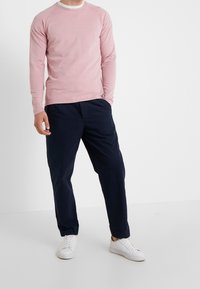 Camicia a maniche lunghe rosa chiaro con scollatura rotonda, abbinata a pantaloni blu scuro e sneakers bianche, che mostrano un design rilassato.