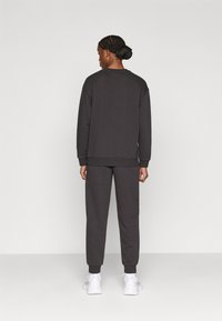 Σετ μπλούζας και παντελονιού σε σκούρο γκρι, κατασκευασμένο από μαλακό ύφασμα. Διαθέτει μακριά μανίκια, ριμπ μανσέτες και άνετη εφαρμογή. Φοριέται με λευκά αθλητικά παπούτσια.