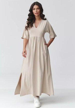 Kobieta o długich ciemnych włosach, ubrana w beżową sukienkę maxi z dekoltem w serek, krótkimi rękawami i bocznymi rozcięciami, w białych trampkach, stoi na białym tle.