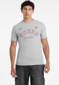 Hall kõrge kvaliteediga hall puuvillane T-särk, millel on punane 'GUESS' logo ja tekst, lühikesed varrukad, ümar kaelus ja kitsas lõige.