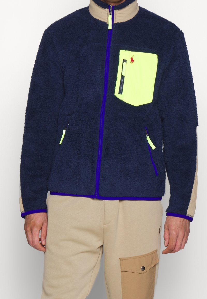 Marinblå fleecejacka med hög krage, kontrasterande neongul ficka och lila dragkedjor. Har sidofickor med dragkedja och beige detaljer.