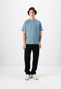 Lys blå bomull t-skjorte med rund hals og korte ermer, kombinert med svarte bukser og beige sneakers; har et lite logo på brystet.