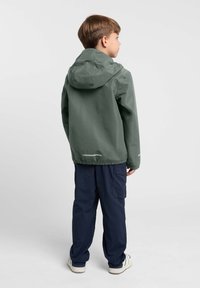 Veste légère verte avec capuche, ourlet élastique et accents réfléchissants, associée à un pantalon cargo bleu marine. Affichant un style décontracté en plein air.