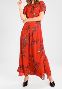 Kvinde iført en rød maxikjole med blomsterprint, korte ærmer og sideslids, parret med sorte spidse hæle.