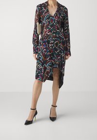 Långärmad klänning med V-ringning i mörk tyg som har ett färgglatt paisley-mönster, hög slits till låret och en lätt svängig silhuett. Kombination med svarta klackar.