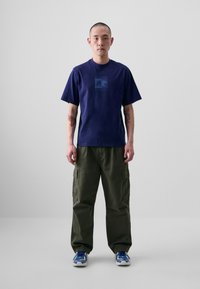T-shirt en coton bleu marine à manches courtes avec un imprimé graphique sur la poitrine, associé à un pantalon cargo vert olive et des baskets bleues.