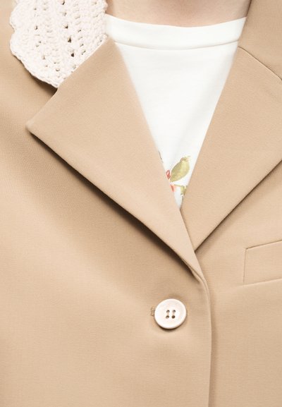 Μπεζ σακάκι με μεγάλους γιακάδες και κούμπωμα με ένα κουμπί, φορεμένο πάνω από άσπρο πουκάμισο με μερική λουλουδάτη εκτύπωση και πλεκτή λεπτομέρεια στον ώμο.