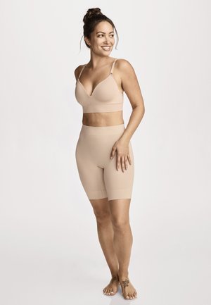 Donna sorridente con chignon che indossa un reggiseno beige senza cuciture e pantaloncini compressivi a vita alta, in piedi a piedi nudi su uno sfondo chiaro.